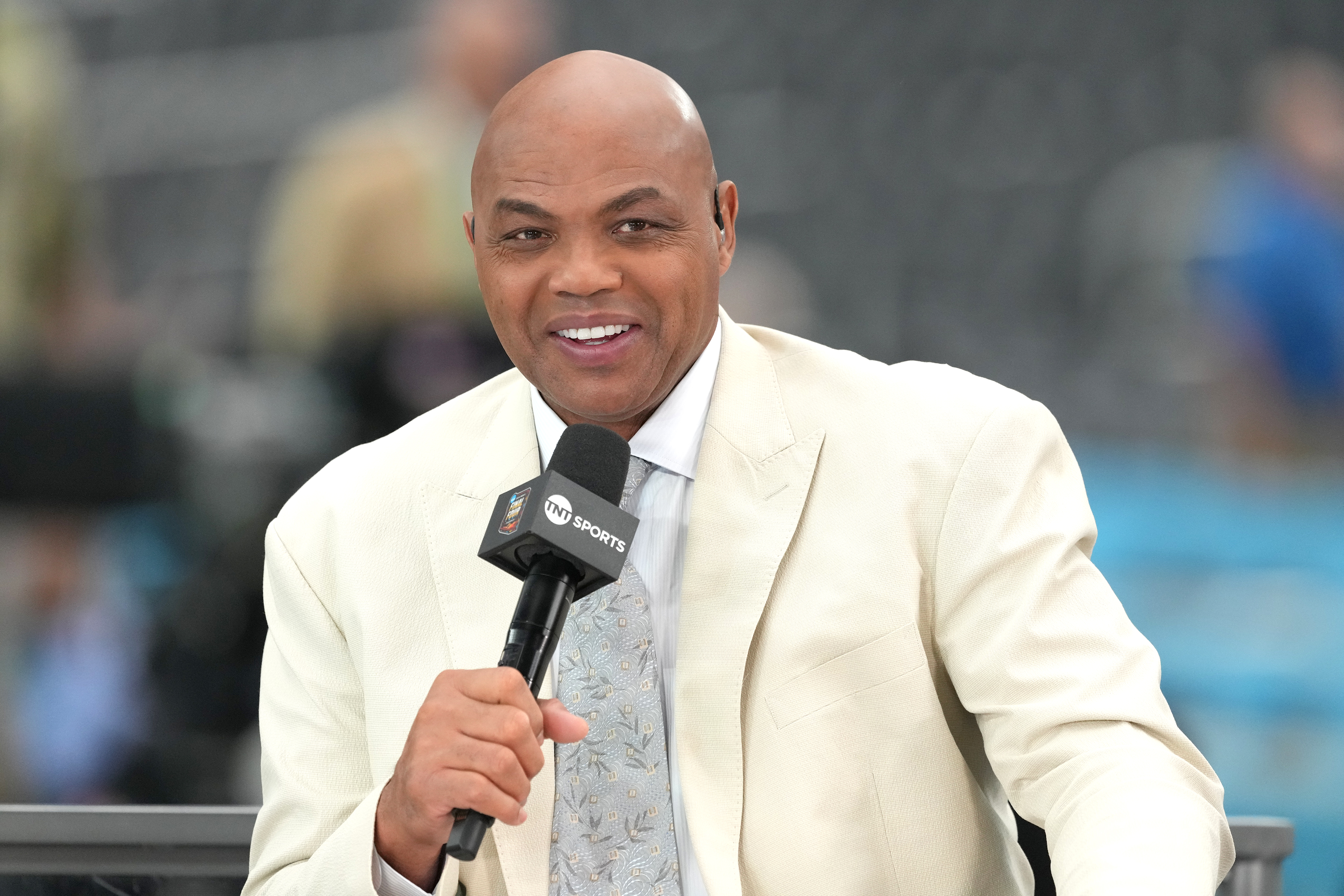 Charles Barkley, wearing a white suit coat and a silver tie, smiling as he speaks into a black microphone in front of a blurred background.