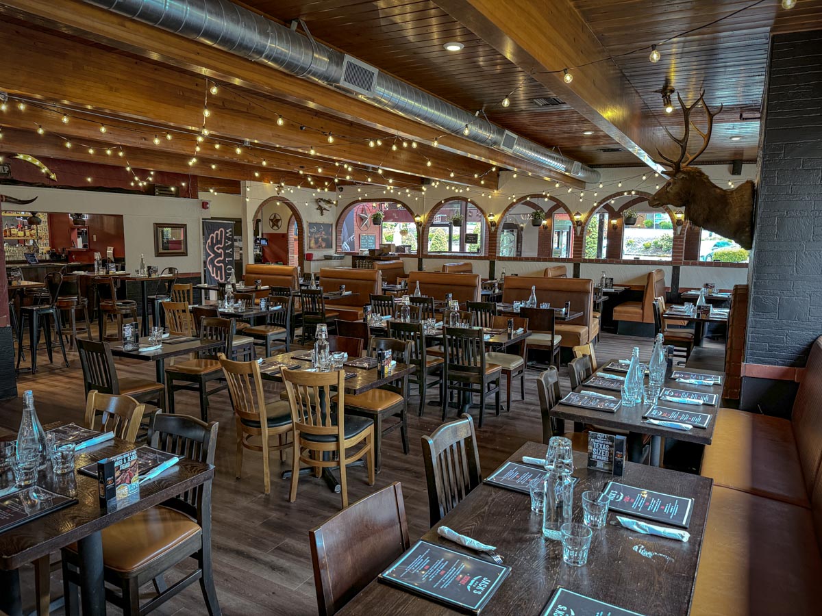 The interior of a large casual restaurant with white walls, wooden tables, and white Christmas lights on the ceiling beams.