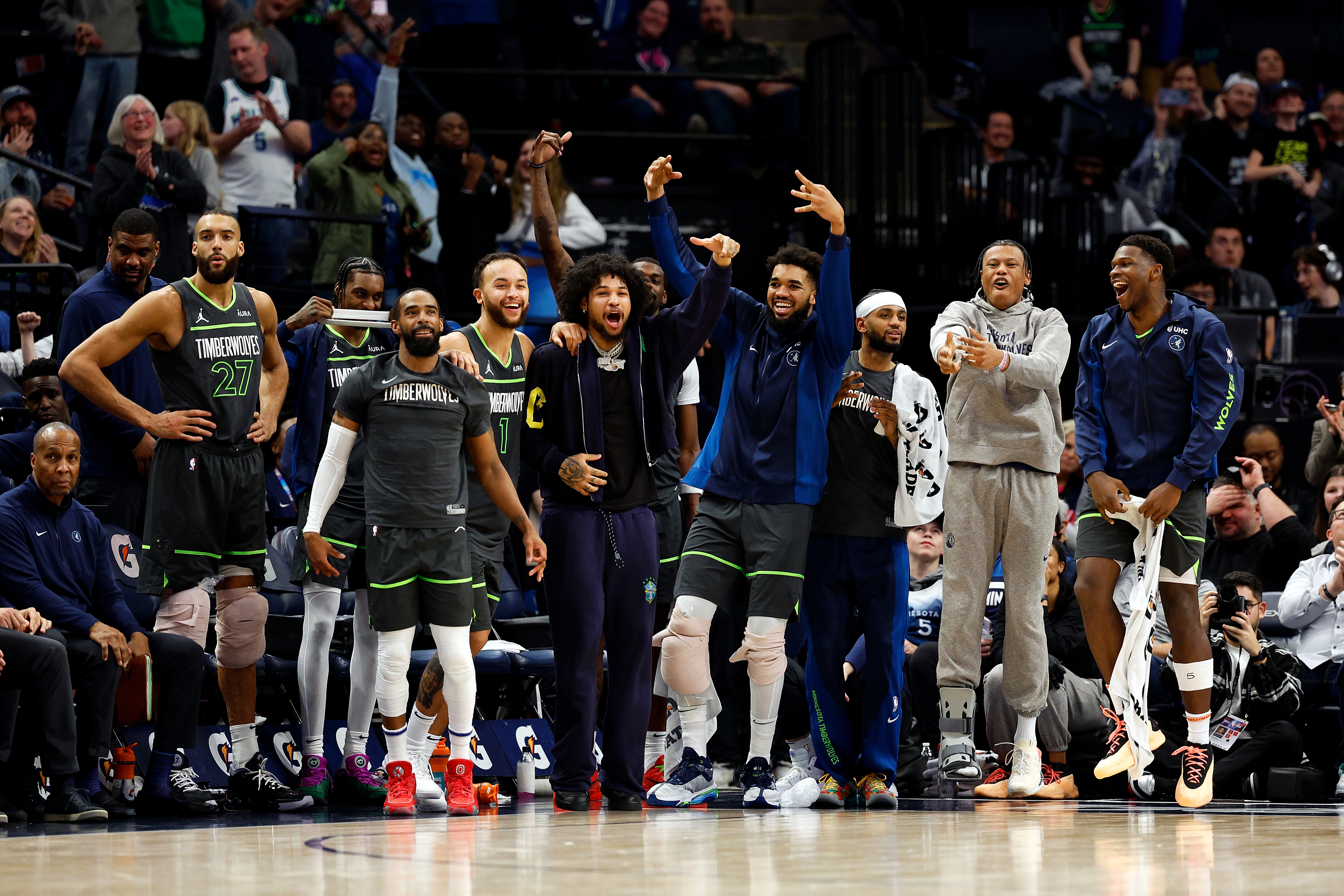 Timberwolves players dressed in uniform standing courtside, smiling and cheering at their teammates on the court.