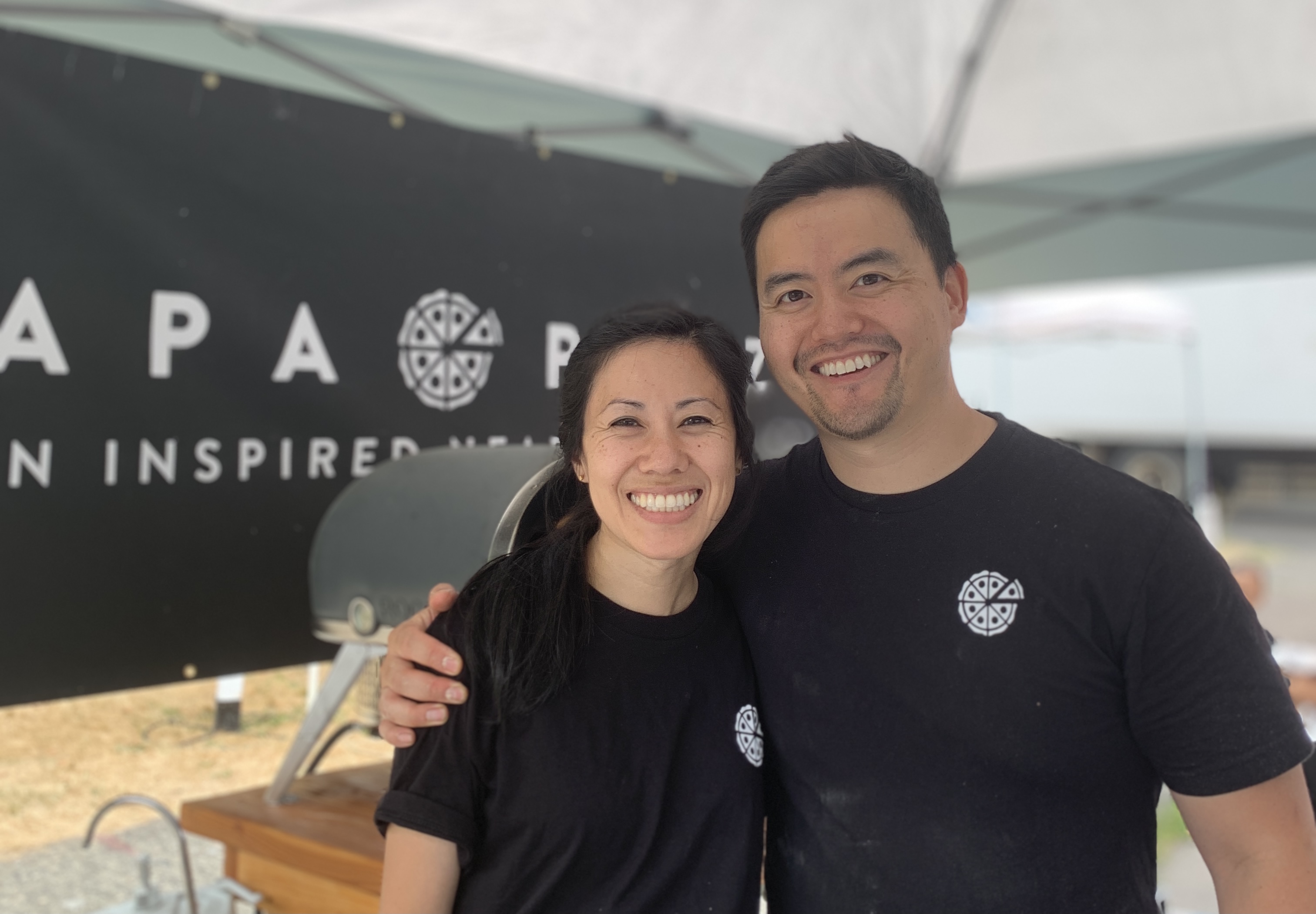 A man and a woman in black shirts stand at a food stall at a farmer’s market. They are the owners of Hapa Pizza.