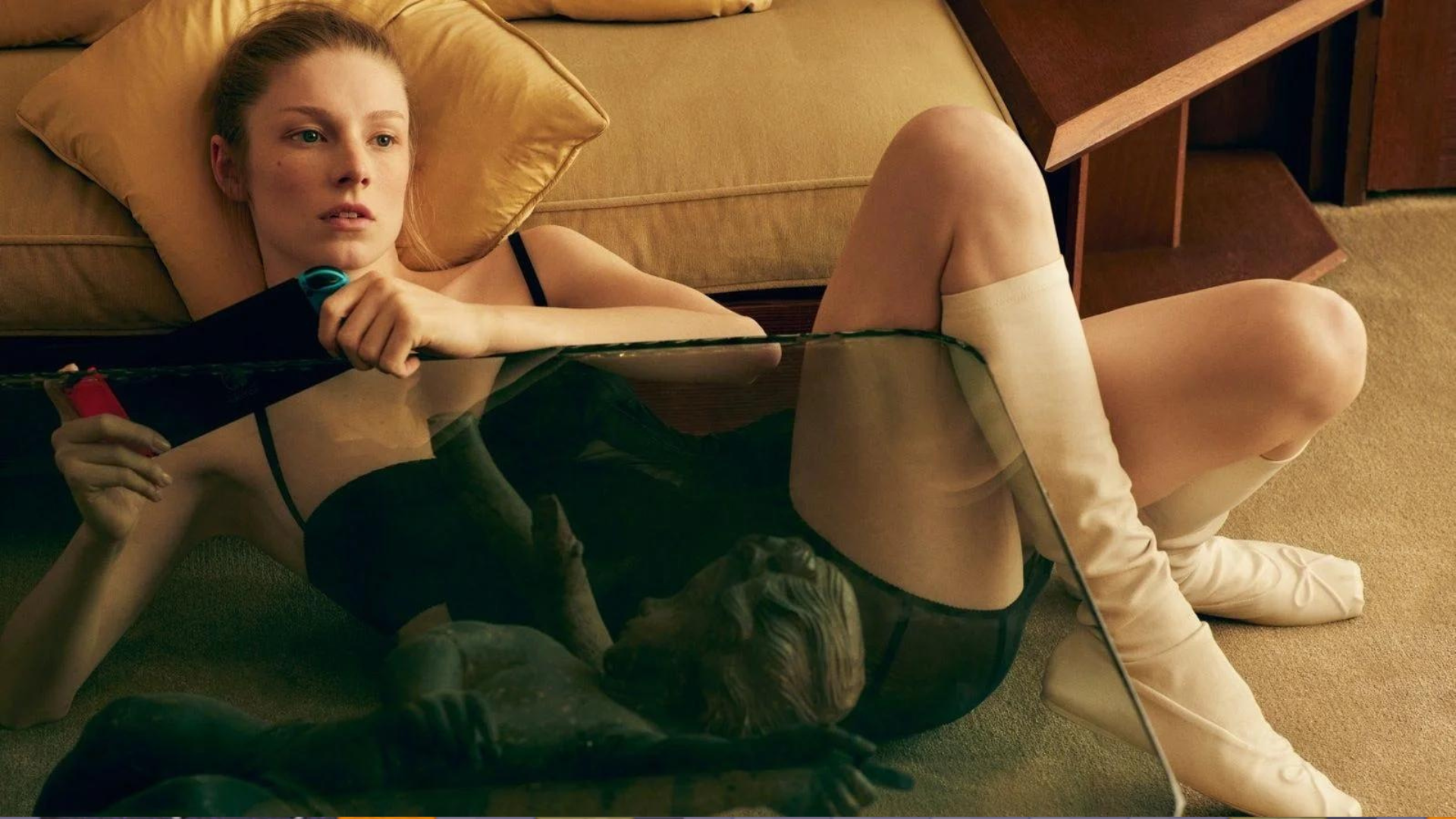 Hunter Schafer lounges on the floor next to a coffee table and couch, her head propped up on a pillow, wearing a black dress and high white boots in a shoot for Vogue Magazine.