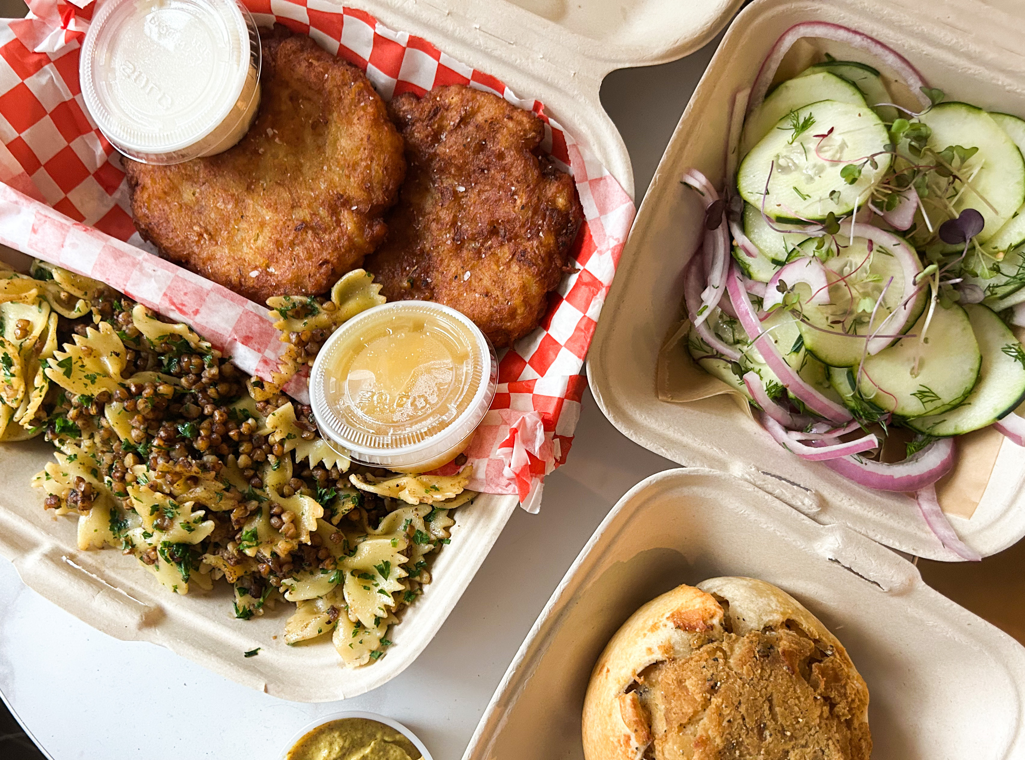 Takeout containers of latkes with sour cream and applesauce, kasha varnishkes, a potato knish, and cucumber salad sit on a table.