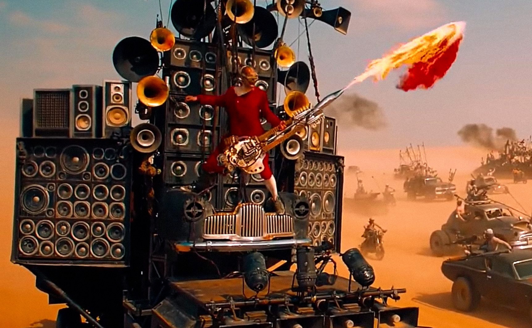 Coma the Doof Warrior, a man in red long johns and a skull mask, blasts fire from an electric guitar while standing atop a vehicle made out of stacked speakers in George Miller’s Mad Max: Fury Road