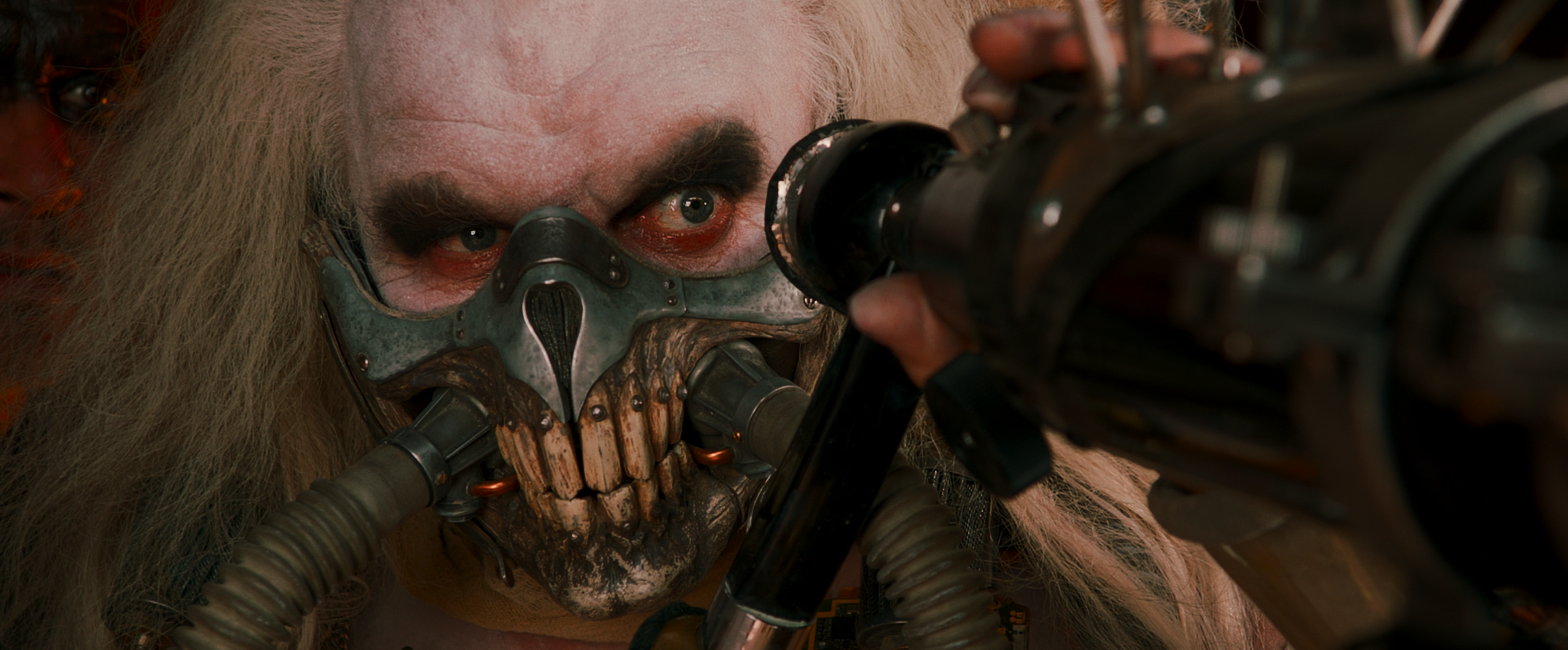 Immortan Joe with white hair red rings under his eyes and a metal cage housing his teeth, looks from behind a telescope