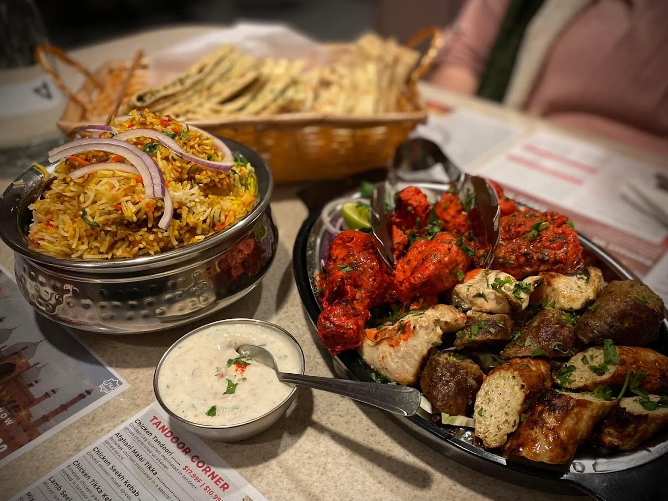 A bread basket with naan, serving bowl of biryani, and platter of tandoori meats.