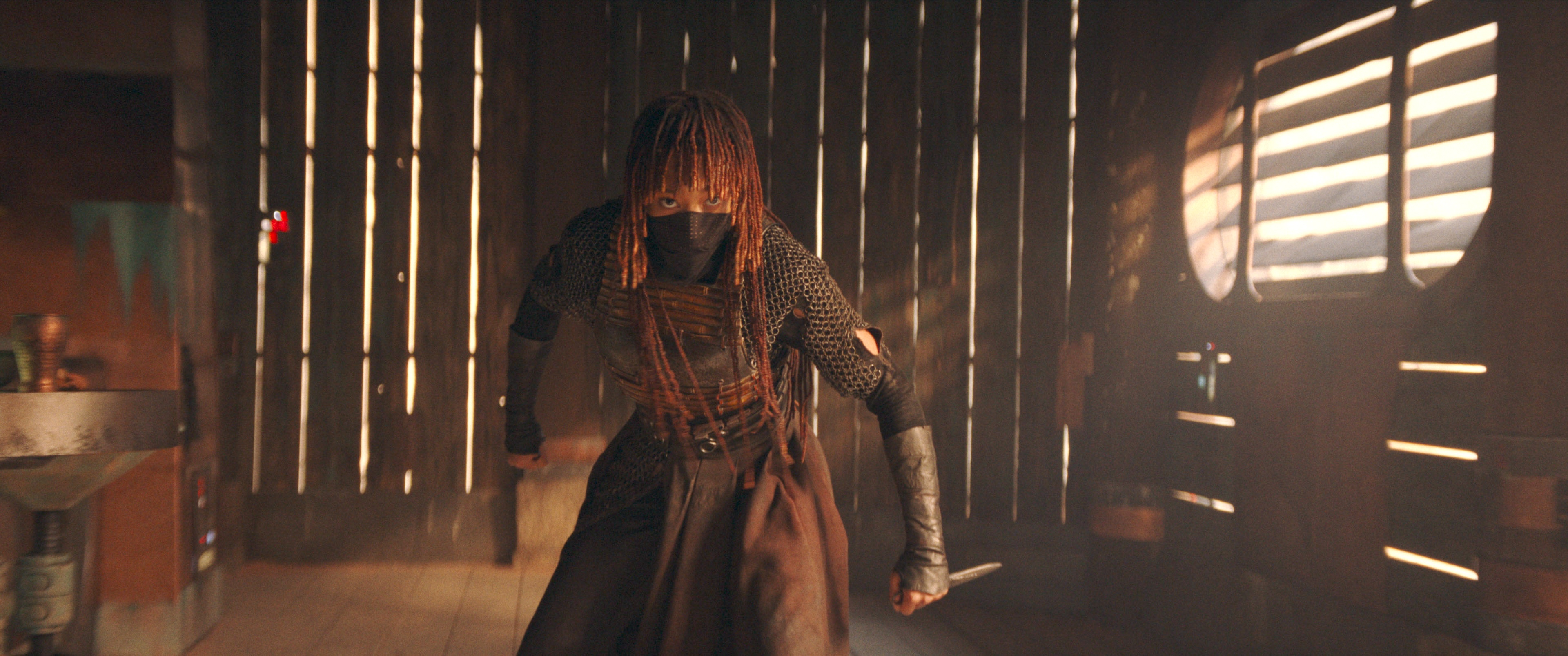 Mae&nbsp;(Amandla Stenberg) in The Acolyte. She’s standing in a fighting stance in a wooden-slatted building, a small dagger gripped in one hand. The bottom of her face is covered by a cloth mask. 