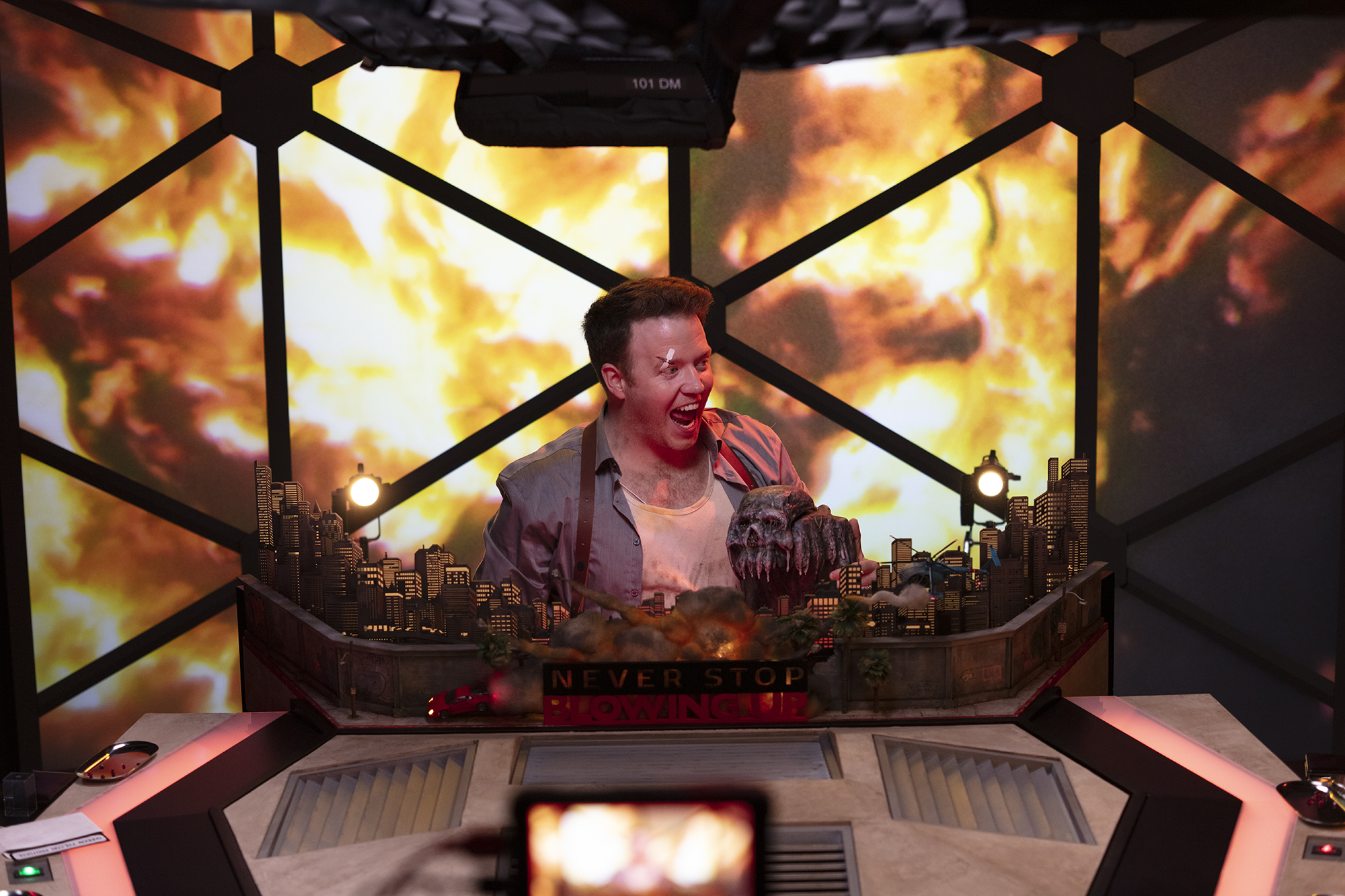 Brennan Lee Mulligan screams in the Game Master seat of the Dimension 20 dome. He’s wearing dirty clothes and has a bandaid on his face, and the screens behind him are showing a huge fiery explosion in Never Stop Blowing Up.