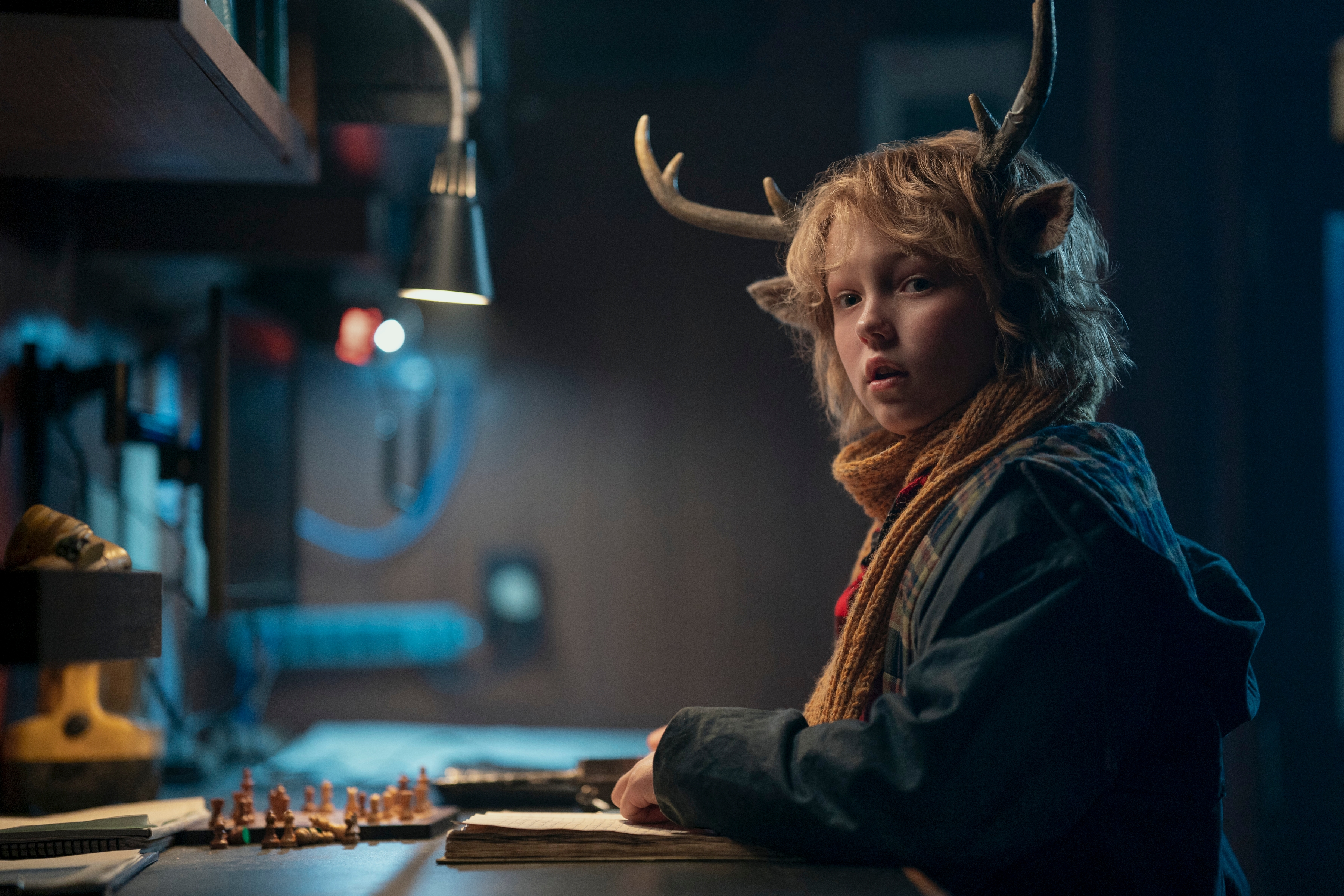 Christian Convery as Gus looks left at the camera while sitting at a desk in Sweet Tooth X3. Gus is a little boy who is dressed warmly, has a shaggy blonde mop, and the ears and antlers of a deer. 