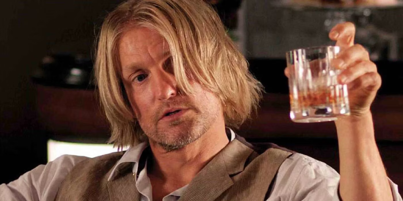 Haymitch, a scruffy blond man played by Woody Harrelson, holds up a glass of whiskey
