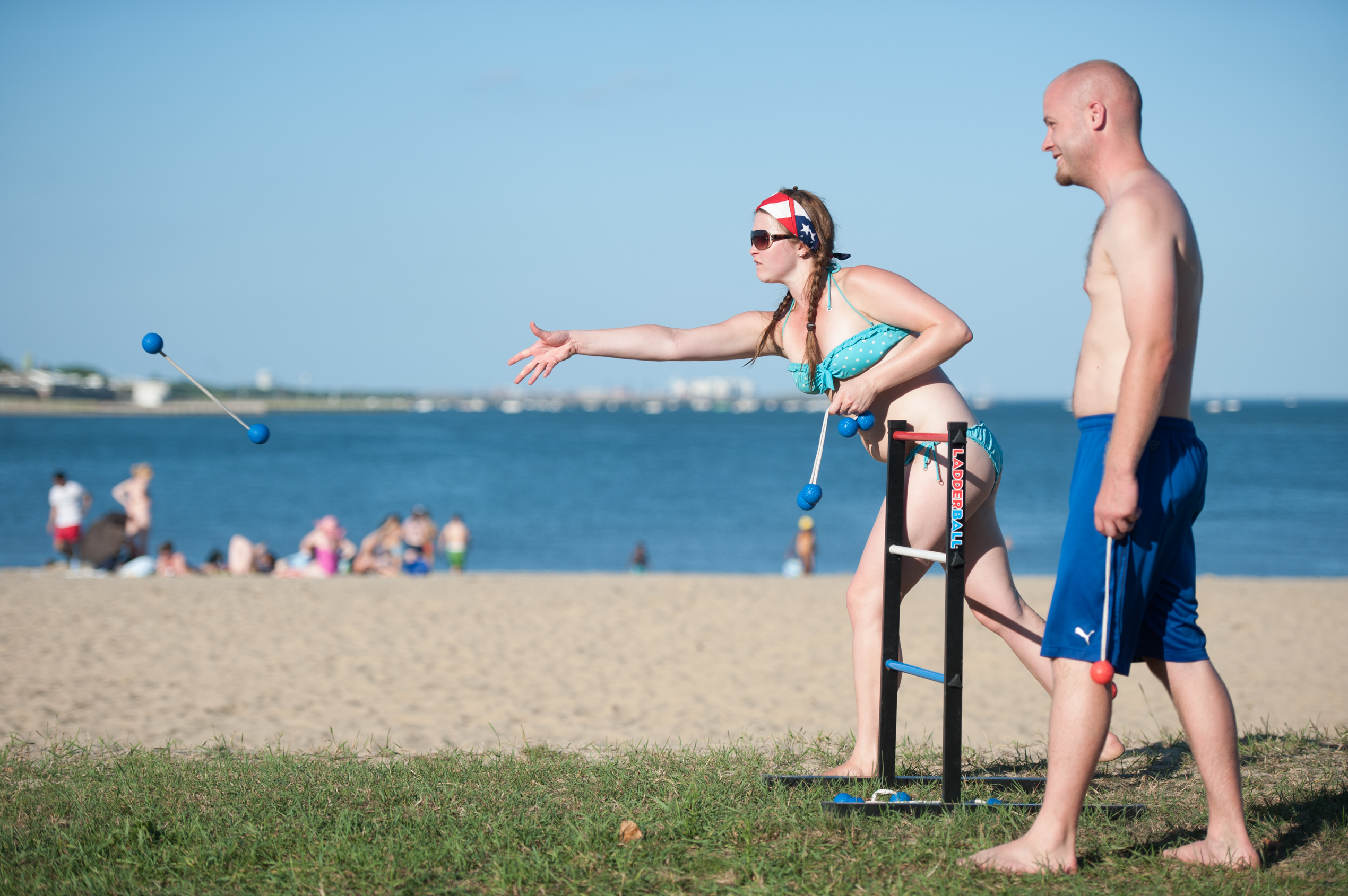 At Carson Beach, in Boston, two beachgoers toss tethered golf balls in a game of Ladderball. The sky is clear, and they’re both wearing swimsuits.