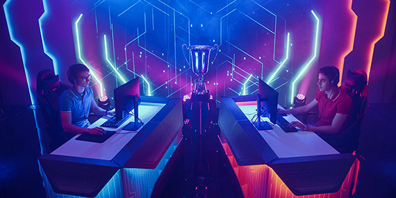 Photograph of two men playing games on computers facing each other.  There are blue and pink vertical lights on either side. 