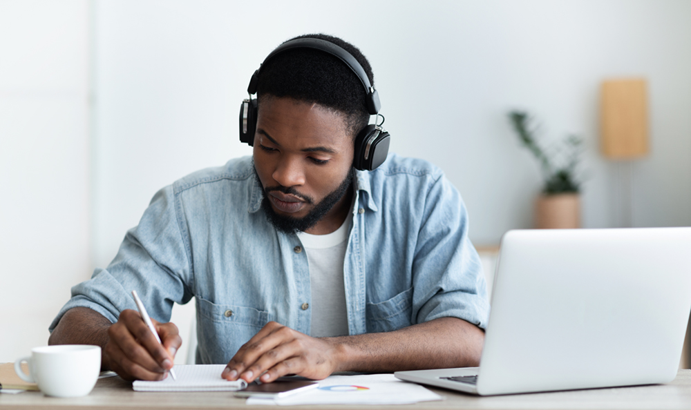 young black man wearing headphones and writing notes sitting at a desk with a laptop
