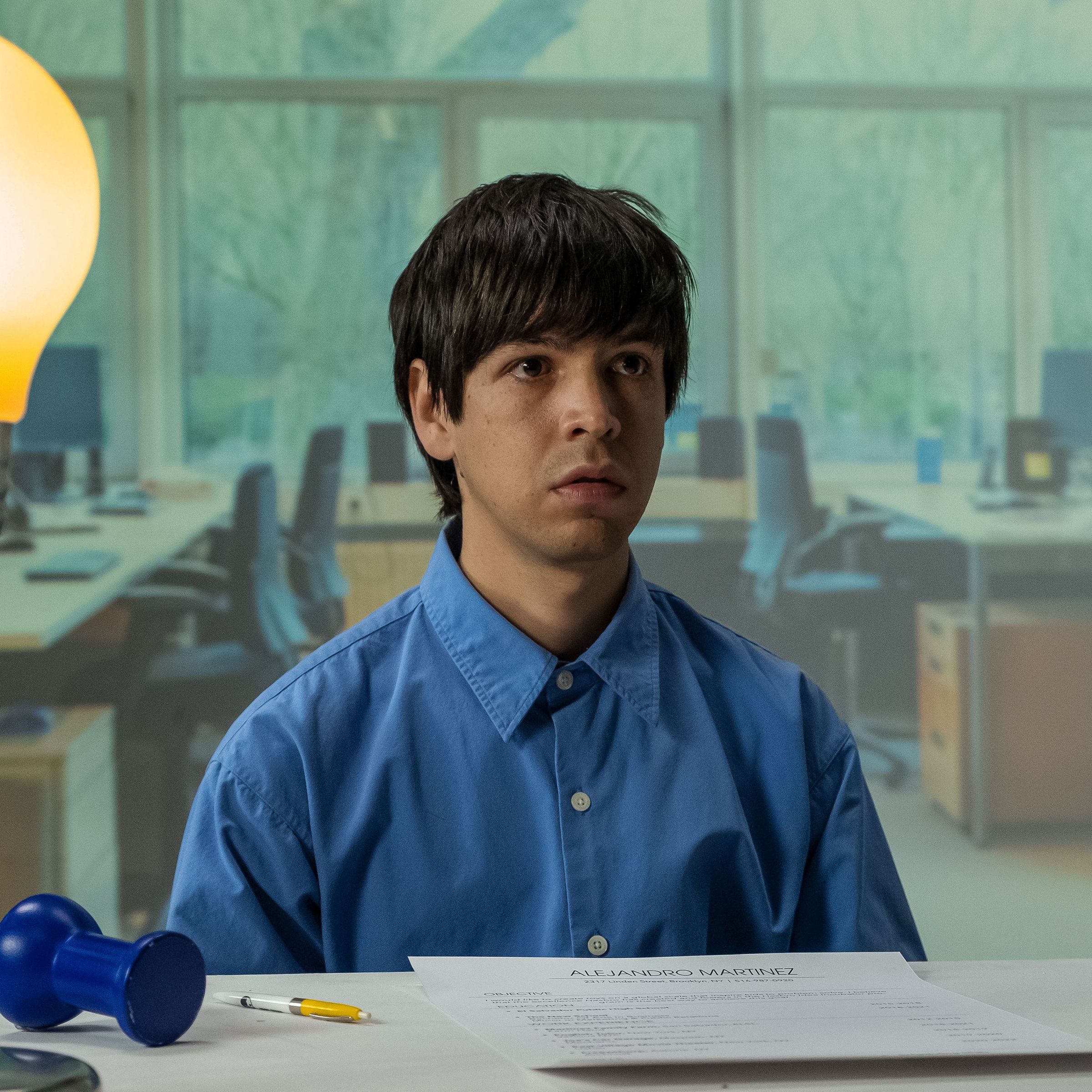 A man in a blue button-down shirt sitting at a desk with a lamp shaped like an oversized light bulb. The man is sitting at a desk on which sits an oversized thumbtack, a pen, and a piece of paper.