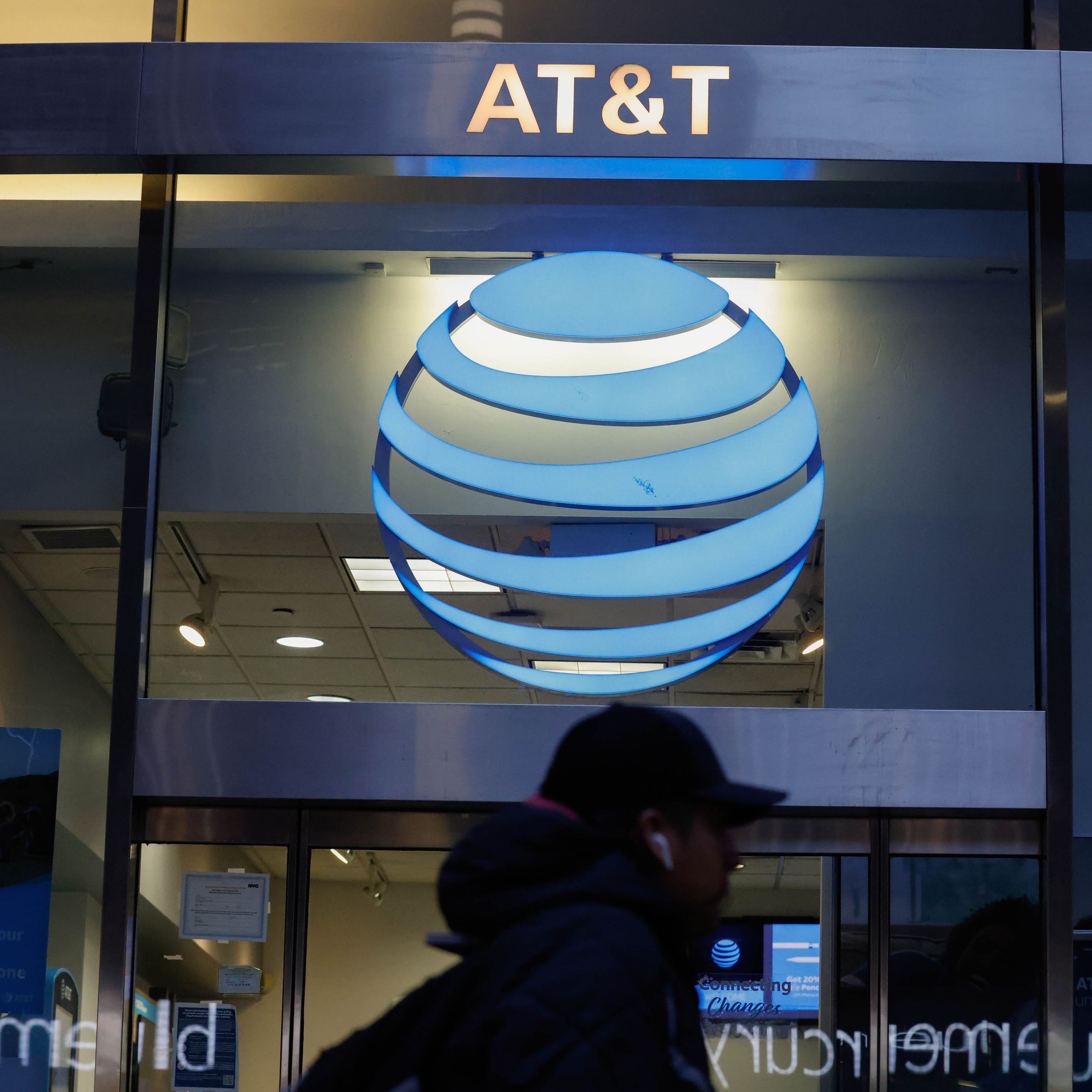 A person wearing earbuds walks past a storefront with the AT&amp;T logo above the person’s head.