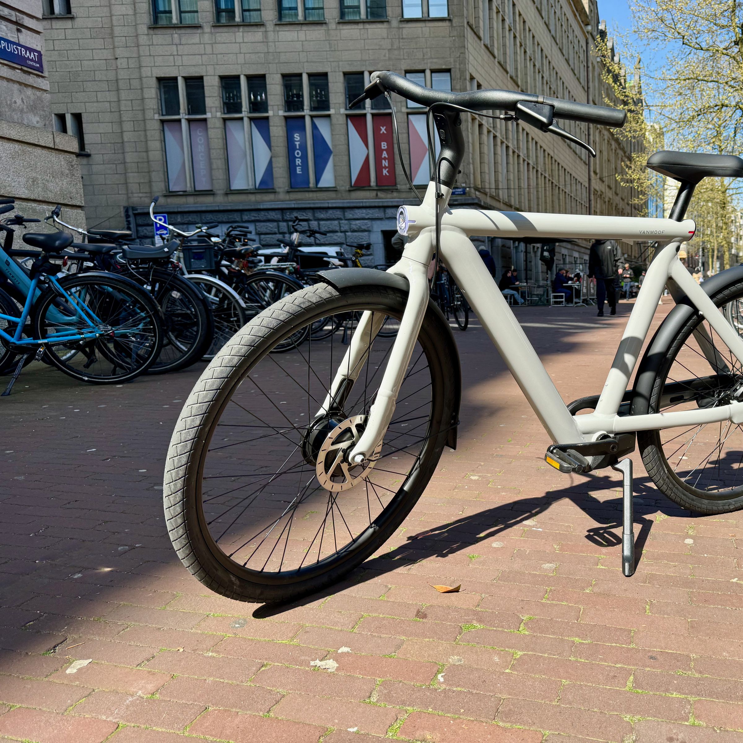 A light grey VanMoof S5 e-bikes sits on a brick sidewalk in Amsterdam between two long rows of bicycles.