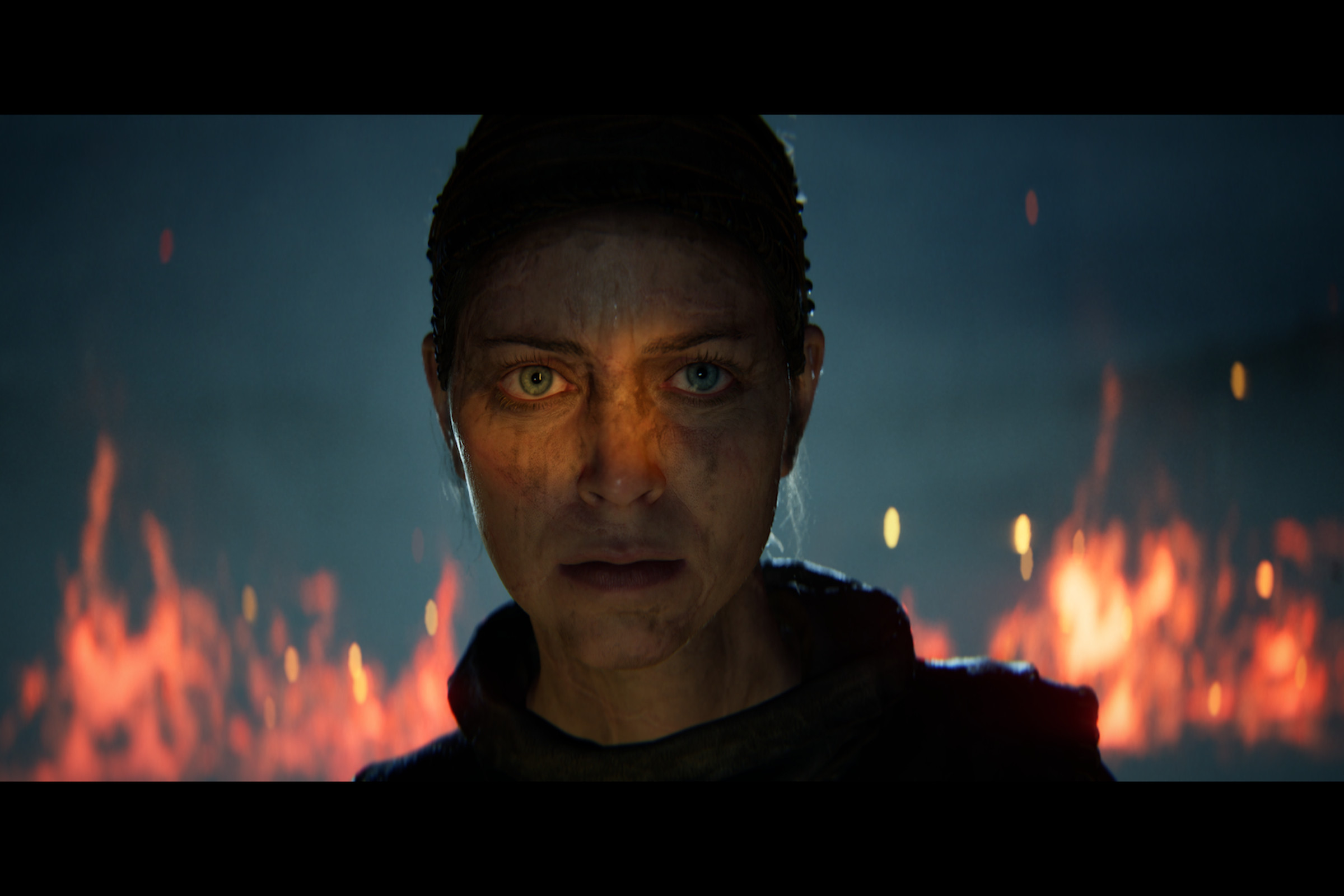 Screenshot from Senua’s Saga: Hellblade II featuring a close up shot of Senua, a woman covered in grime with flames burning behind her.