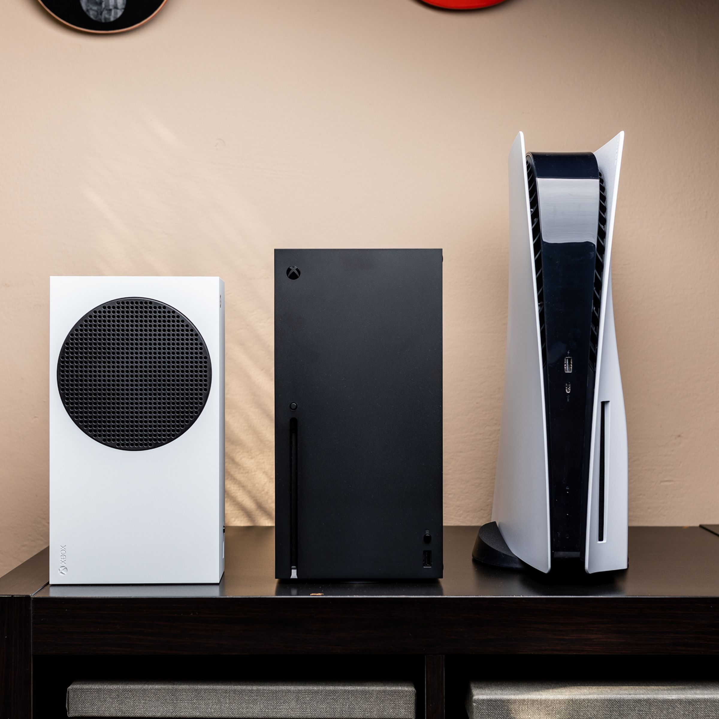 Left to right: the Xbox Series S, Xbox Series X, and PlayStation 5 consoles standing vertically on a TV cabinet.
