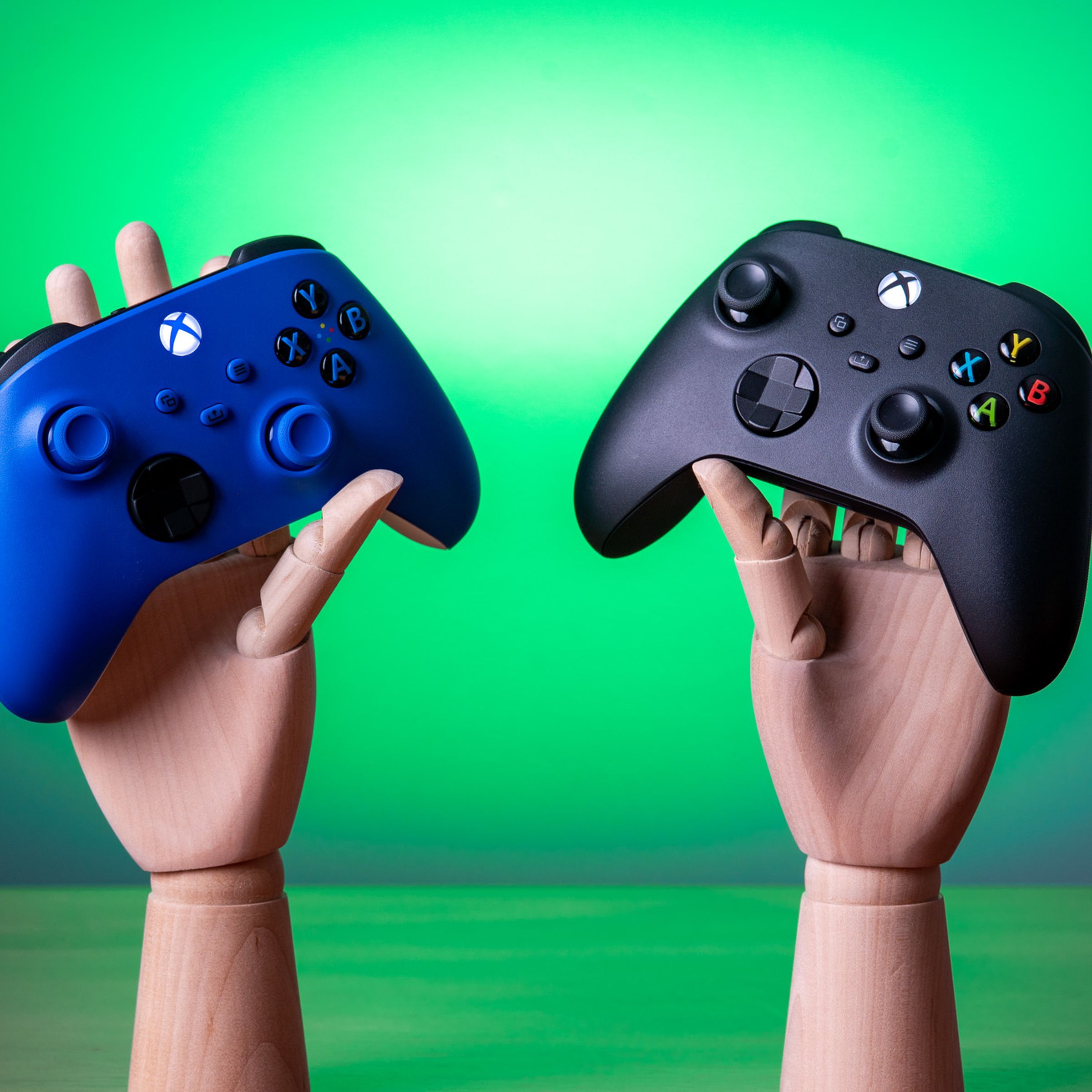 Two Xbox controllers, one blue and one black, being held by wooden mannequin hands.