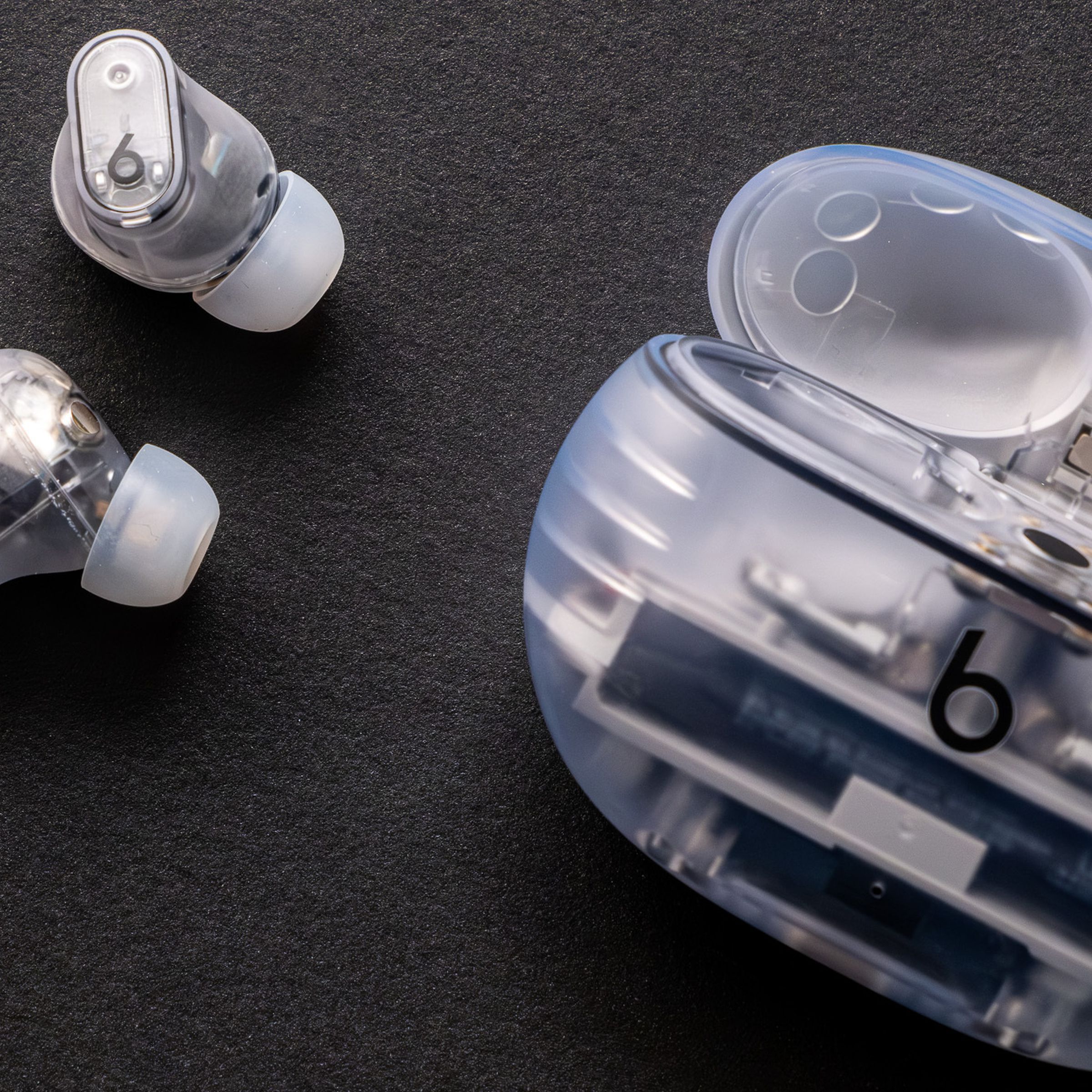 The see-through Beats Studio Buds Plus and their matching charging case, against a black background.