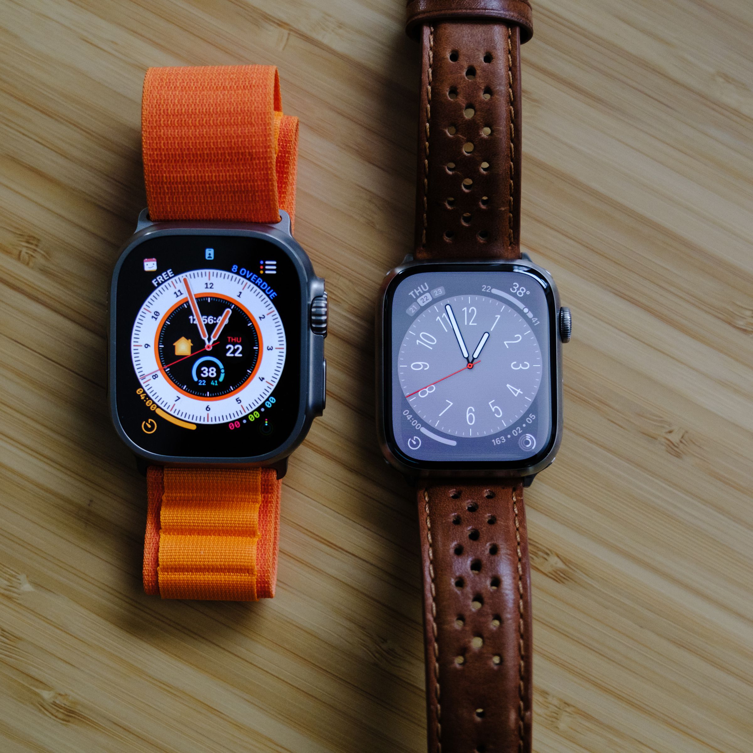 An Apple Watch Ultra next to a Series 7 Edition on a light wood table, viewed from the top.