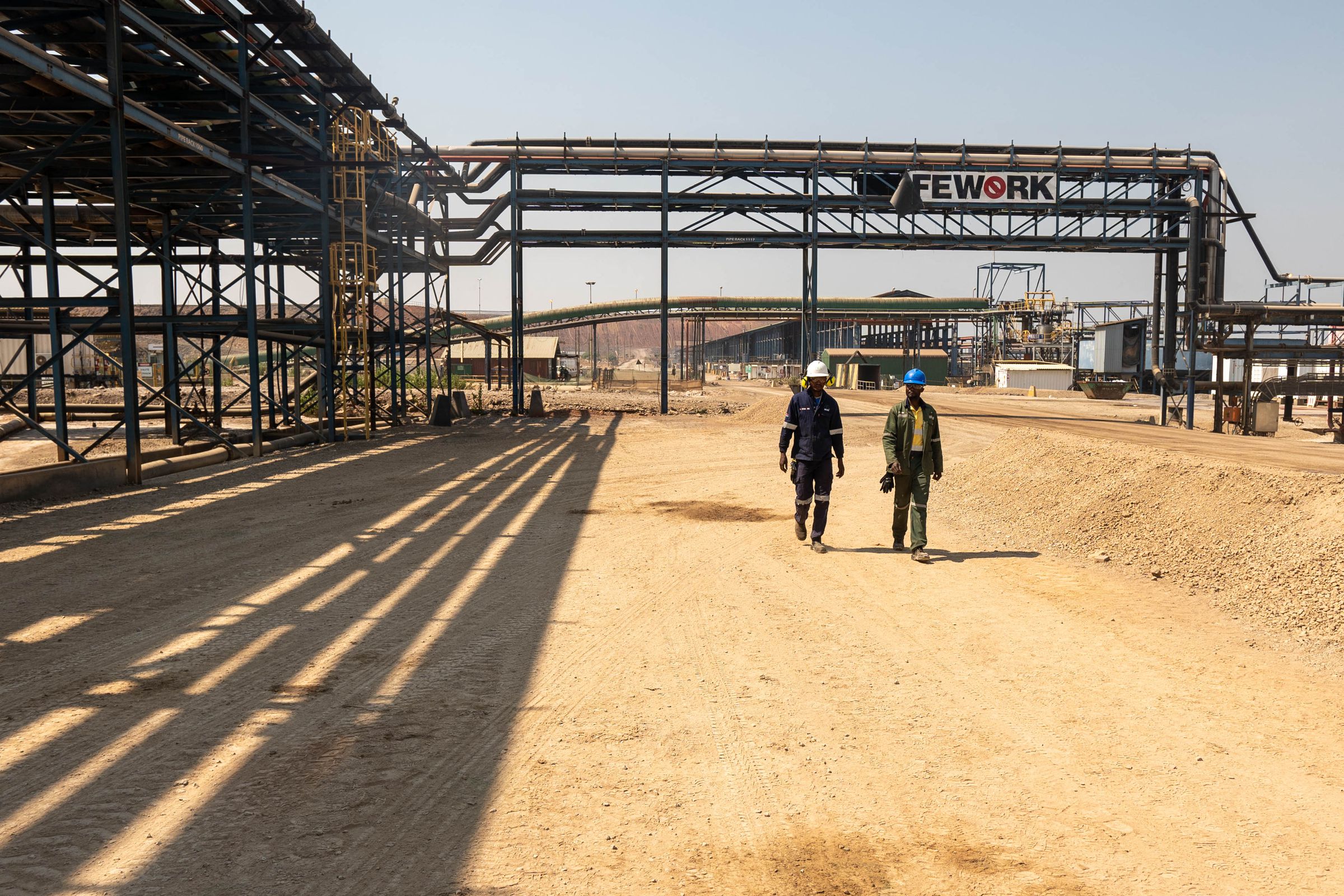 Two workers wearing hard hats walk through an industrial area.