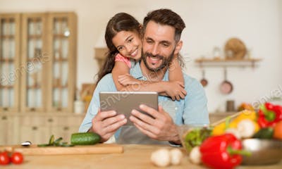 Happy father and his daughter using digital tablet, girl embracing dad from back while cooking