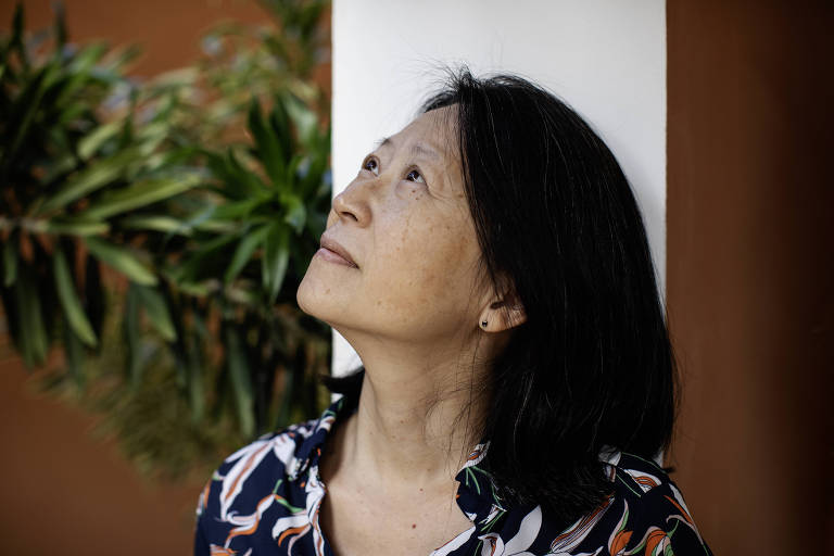 Uma mulher asiática com expressão pensativa olha para cima, talvez contemplando algo fora do quadro. Ela está ao ar livre, com uma planta ao fundo, sugerindo um momento de conexão com a natureza ou introspecção.
