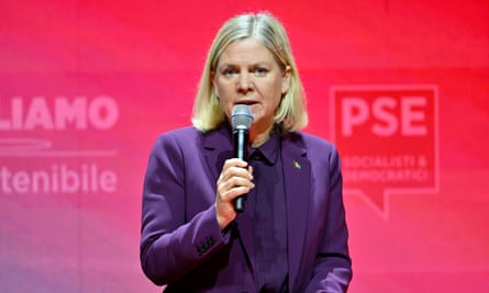 Magdalena Andersson speaks holding a microphone on a stage with a red backdrop