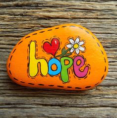 an orange rock with the word hope painted on it and flowers in the center, sitting on a wooden surface