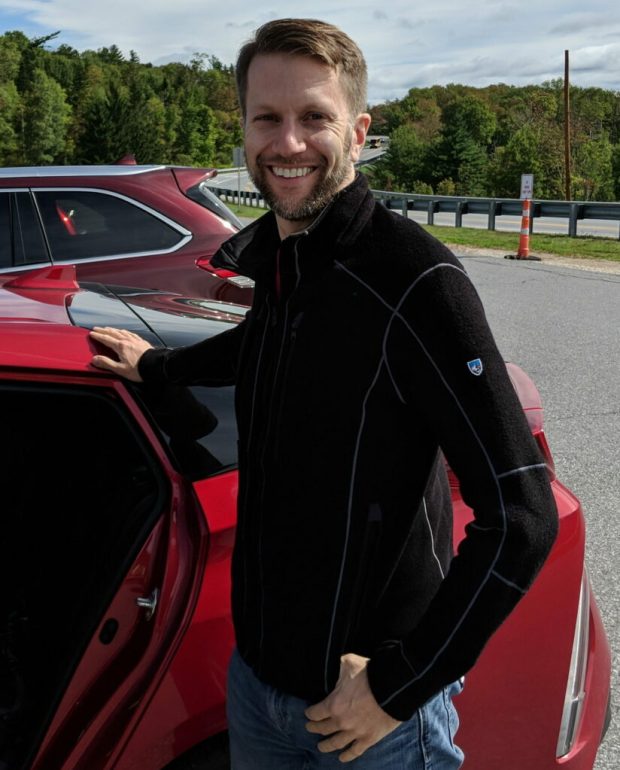 Portrait profile photo of smiling, bearded man standing next to red car