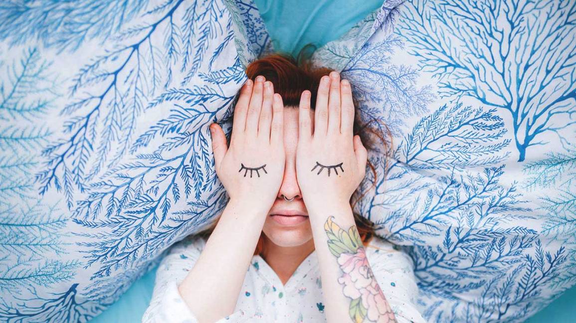 Woman in bed covering her eyes with "closed eyelids" drawn on the backs of her hands