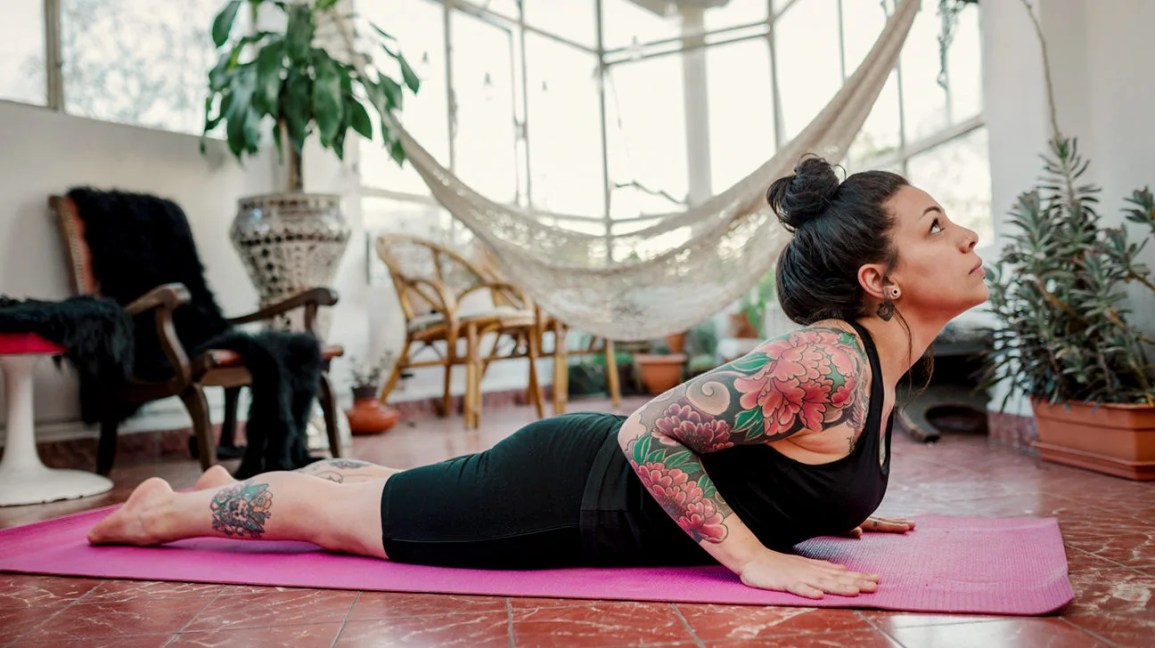 A woman lying on her stomach on a yoga mat in her living room, doing a cobra pose. 