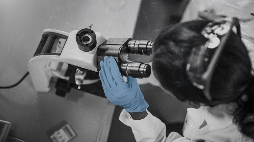 a person wearing a white coat and blue gloves is looking into a microscope
