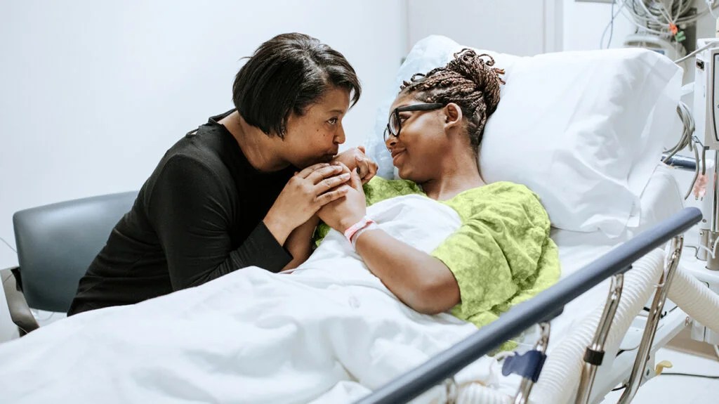 A woman in a hospital bed, holding hands with a relative sat at her side