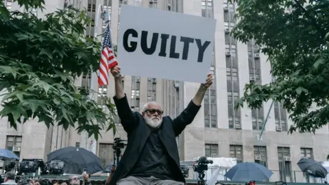 A man holds a sign reading "Guilty" and an American flag outside court