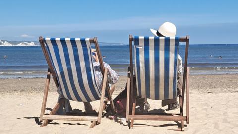 Two people in deck chairs on a sunny beach looking out to sea, seen from behind