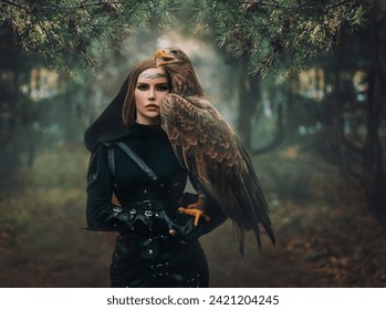 art portrait real people fantasy woman holding white-tailed eagle wild bird on hand. Elf warrior girl beauty face with brown bird walking in forest. Lady queen hunter black costume hood dark trees fog
