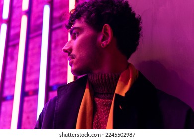 Low angle of young male with curly hair standing against wall near glowing pink neon lights looking away Arkivfotografi