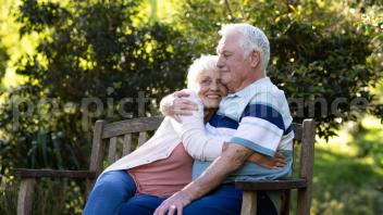 Happy caucasian senior couple sitting on bench and embracing in sunny garden, copy space