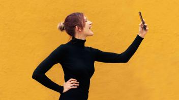 Young woman with mouth open taking selfie standing by yellow wall model released, Symbolfoto, LMCF01050