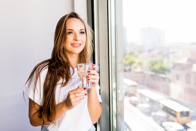Bezpłatne zdjęcie młoda piękna dziewczyna lub kobieta ze szklanką wody w pobliżu okna w białej koszuli i szarej szacie