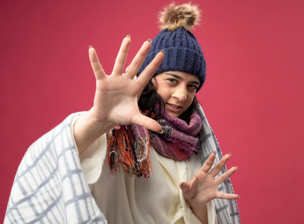 Bezpłatne zdjęcie niezadowolona młoda chora kobieta ubrana w szatę zimową czapkę i szalik owinięty w kratę patrząc na przód wyciągając ręce z tynkiem na nosie odizolowanym na różowej ścianie