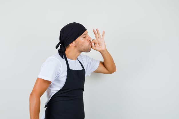Bezpłatne zdjęcie piekarz w koszulce, fartuch pokazuje pyszny gest całując palce i wyglądając na zachwyconego