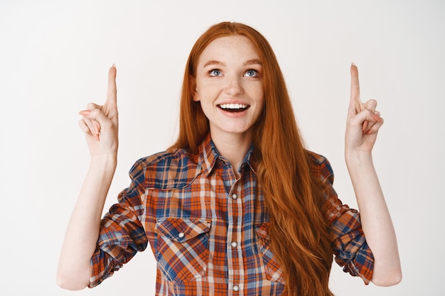 Bezpłatne zdjęcie piękna nastolatka z rudymi długimi włosami, patrząca i wskazująca palce w górę zdumiona, stojąca nad białą ścianą