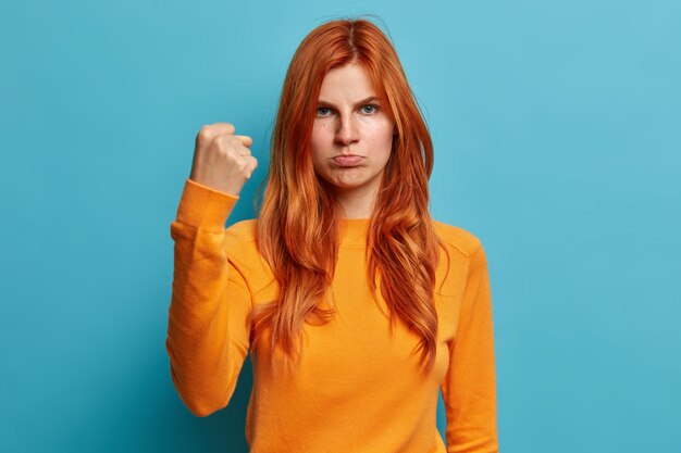 Poważna rudowłosa Europejka wygląda gniewnie i pokazuje pięść, prosząc, by nie zawracać jej warg w torebce, i ma zirytowany wyraz twarzy ubrany w swobodny pomarańczowy sweter.