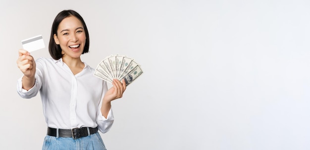 Bezpłatne zdjęcie szczęśliwa koreańska kobieta trzymająca kartę kredytową i pieniądze dolary uśmiechnięta i śmiejąca się na tle białego studia