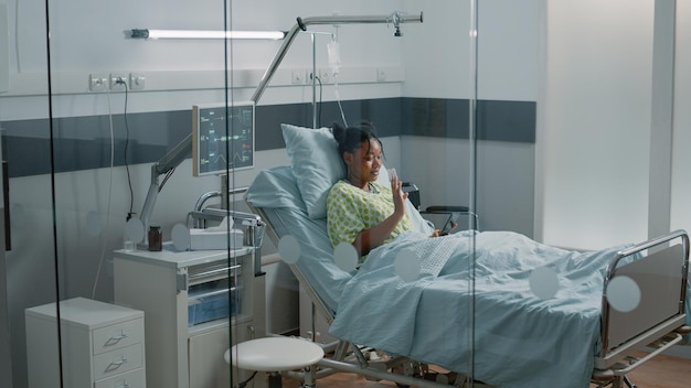 Foto grátis jovem mulher acenando para uma videochamada em smartphone na cama da enfermaria de hospital. paciente com bolsa de gotejamento iv e oxímetro usando conferência remota online na internet para falar com amigos e se recuperando