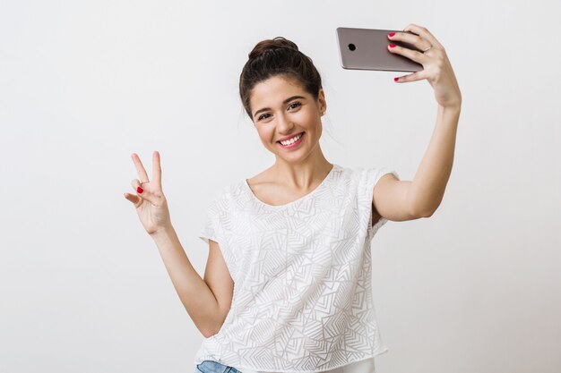 Foto grátis jovem atraente mulher sorridente com blusa branca segurando um telefone inteligente, mostrando o símbolo da paz, emoção positiva, fazendo selfie foto,