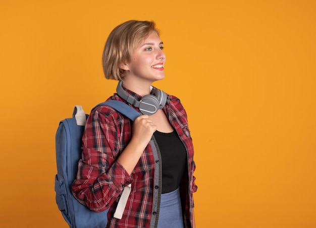Foto grátis jovem estudante eslava sorridente com fones de ouvido e mochila parada de lado