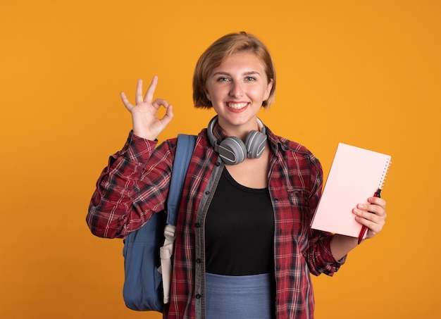 Foto grátis jovem estudante eslava sorridente com fones de ouvido usando uma mochila segurando um caderno e uma caneta fazendo gestos com a mão bem